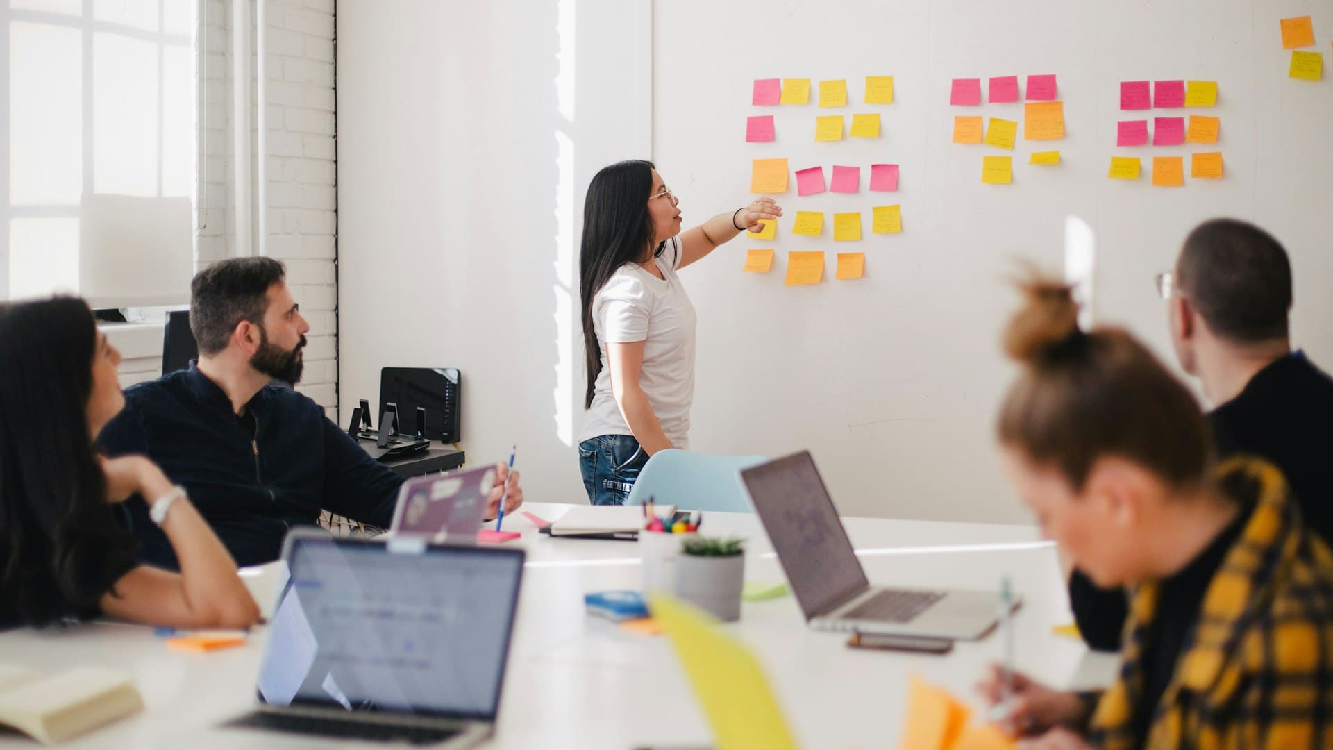 Modern team collaborating around a table with laptops and sticky notes in a bright office workspace