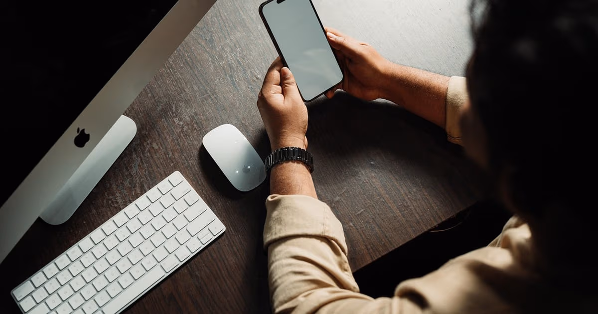 Person at a desk using a smartphone next to a desktop computer, representing Instagram automation tools for small businesses