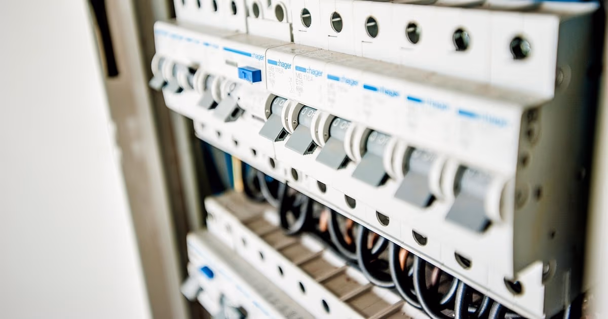 Electrician at an open distribution panel checking estimates and permit tasks on a phone, representing a CRM built for electrical contractors